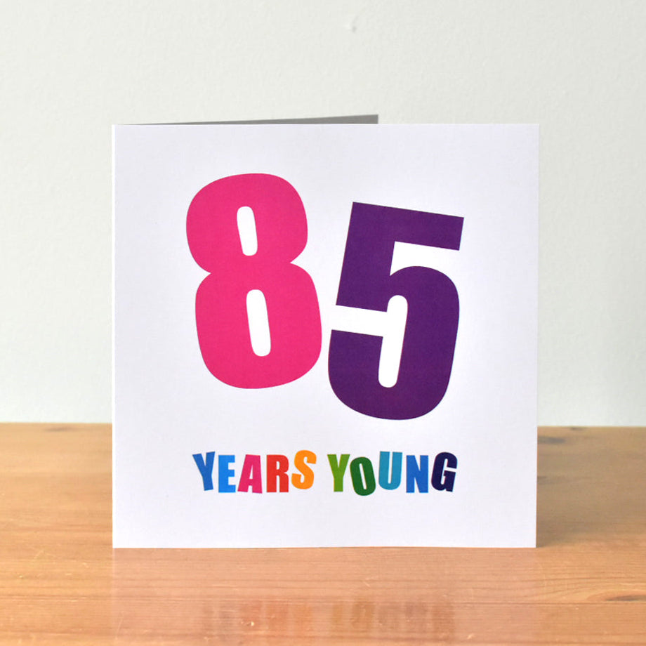 Square white birthday card featuring the large, bold number "85." The "8" and "5" are each at a different angle and rendered in unique bold colours of bright pink and purple. Below this, the phrase "Years Young" appears, with every letter in its own bold colour.