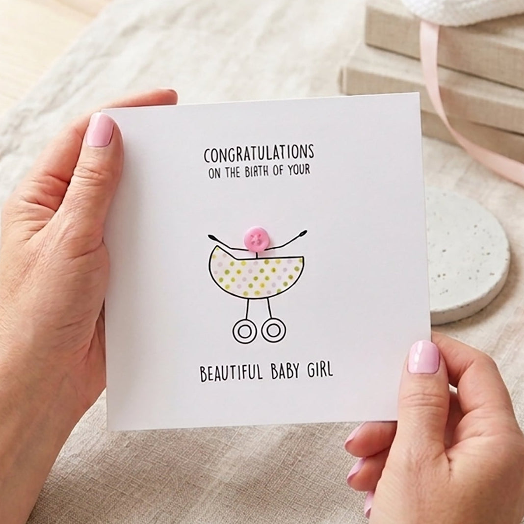 Person holding a baby shower card with a baby girl design on a table with books and a vase.