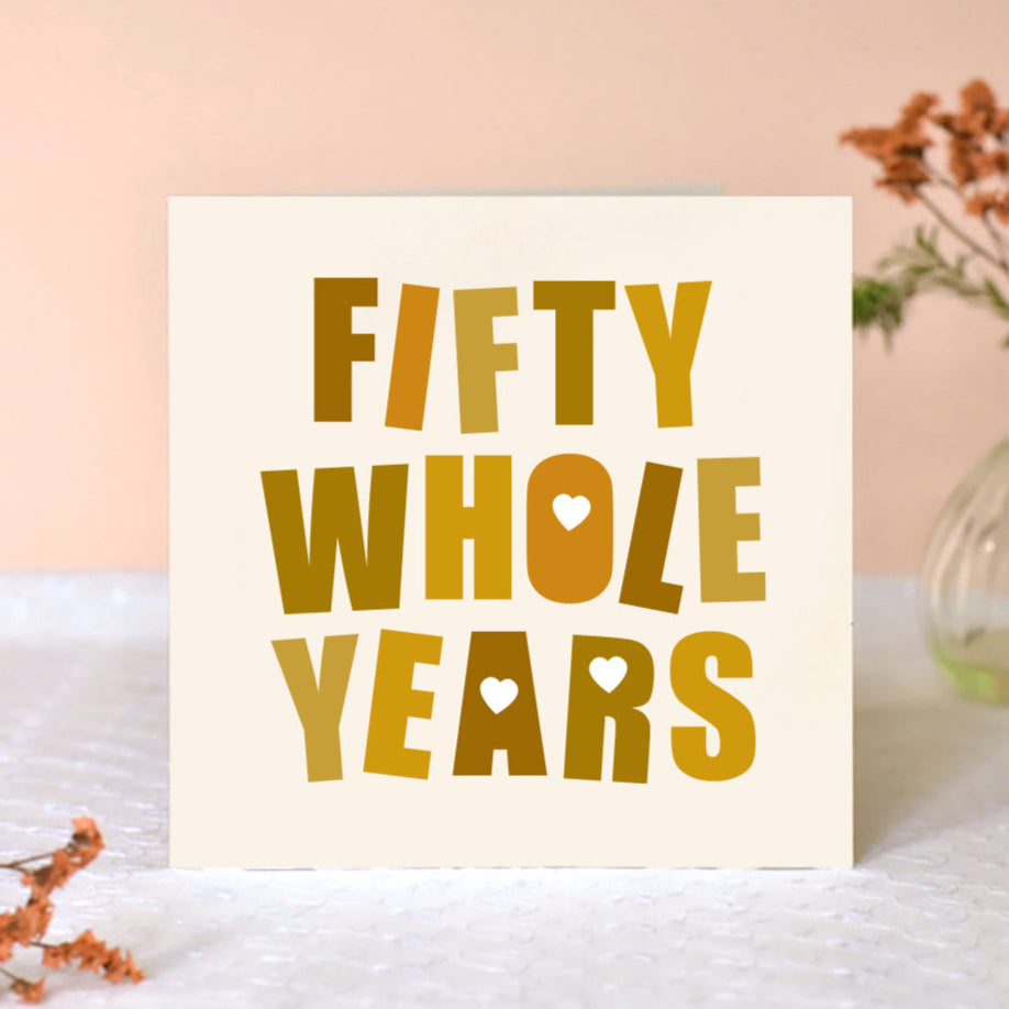 Cream square card with the words Fifty Whole Years in big bold font in shades of gold. Some of the letters are punched with love hearts.
