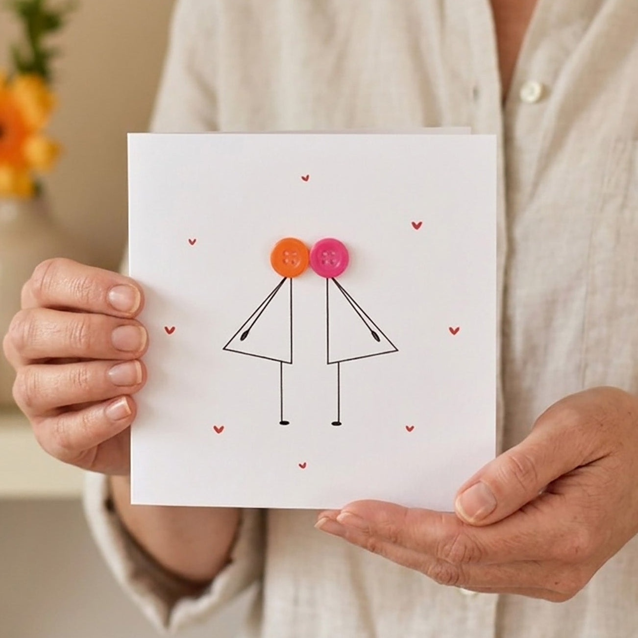 Person holding a card with a card design of two female stick characters with colourful buttons. kissing each other.