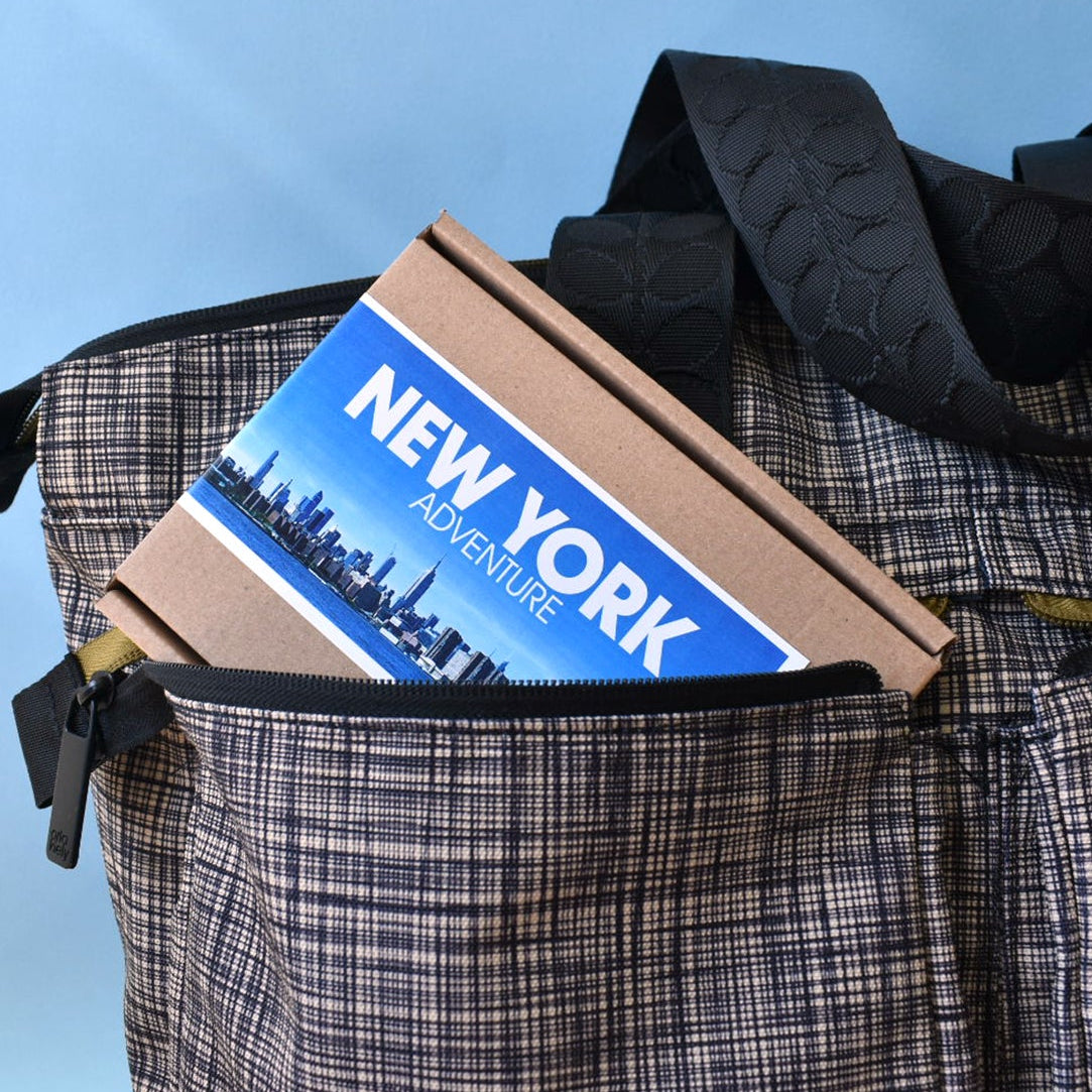 A close-up of a plaid tote bag with black textured shoulder straps, shown holding a brown paperboard box labeled 'NEW YORK ADVENTURE'.