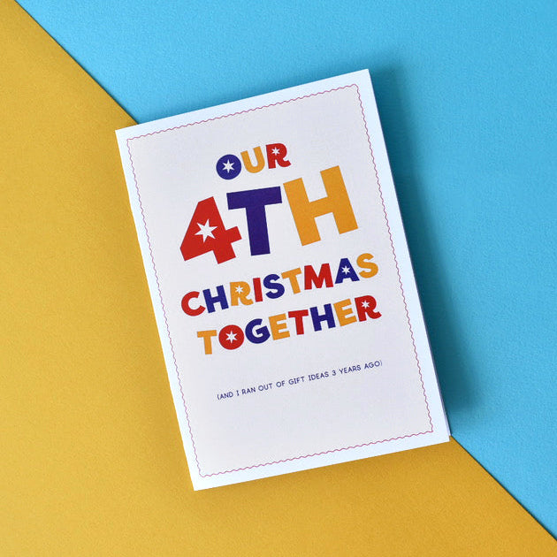 A square greeting card with the text 'OUR 4TH CHRISTMAS TOGETHER' in colorful lettering, placed on a diagonally split yellow and blue background.