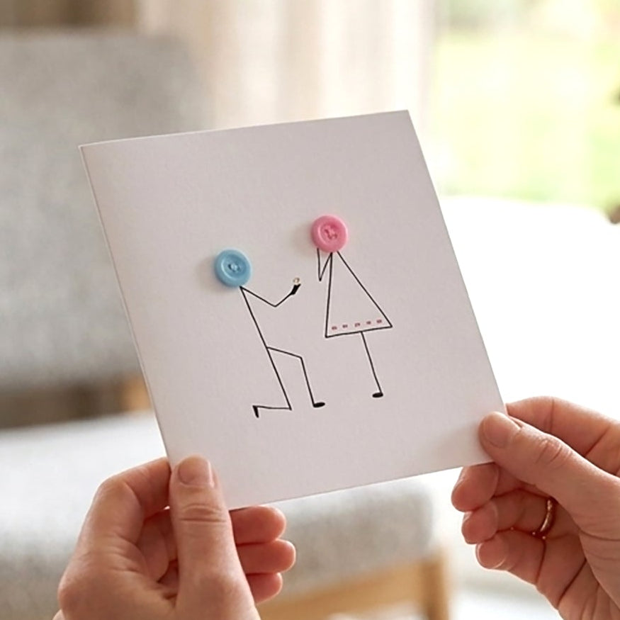 Close-up shot from behind a young woman in a cozy cream jumper holding a handmade stick-figure proposal card inside a neutral, modern living room.