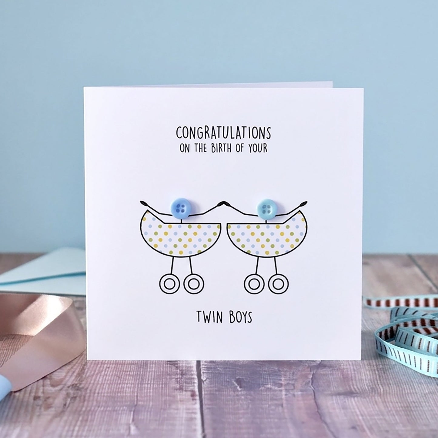A white square greeting card standing on a wooden surface, printed with the message 'CONGRATULATIONS ON THE BIRTH OF YOUR Twin Boys' and a simple line drawing of two baby prams with blue balloons. The card is styled with a pale blue background, ribbons, and a measuring tape.