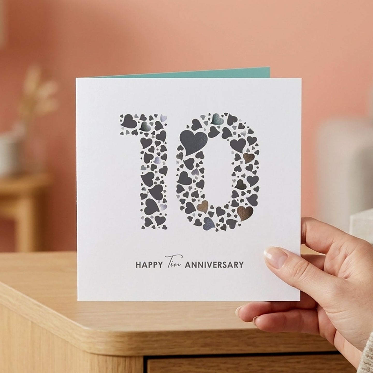 A close-up lifestyle shot of a person's hand holding a square white 10th tin wedding anniversary card featuring a love heart typography design, set against a blurred living room background with soft coral walls.