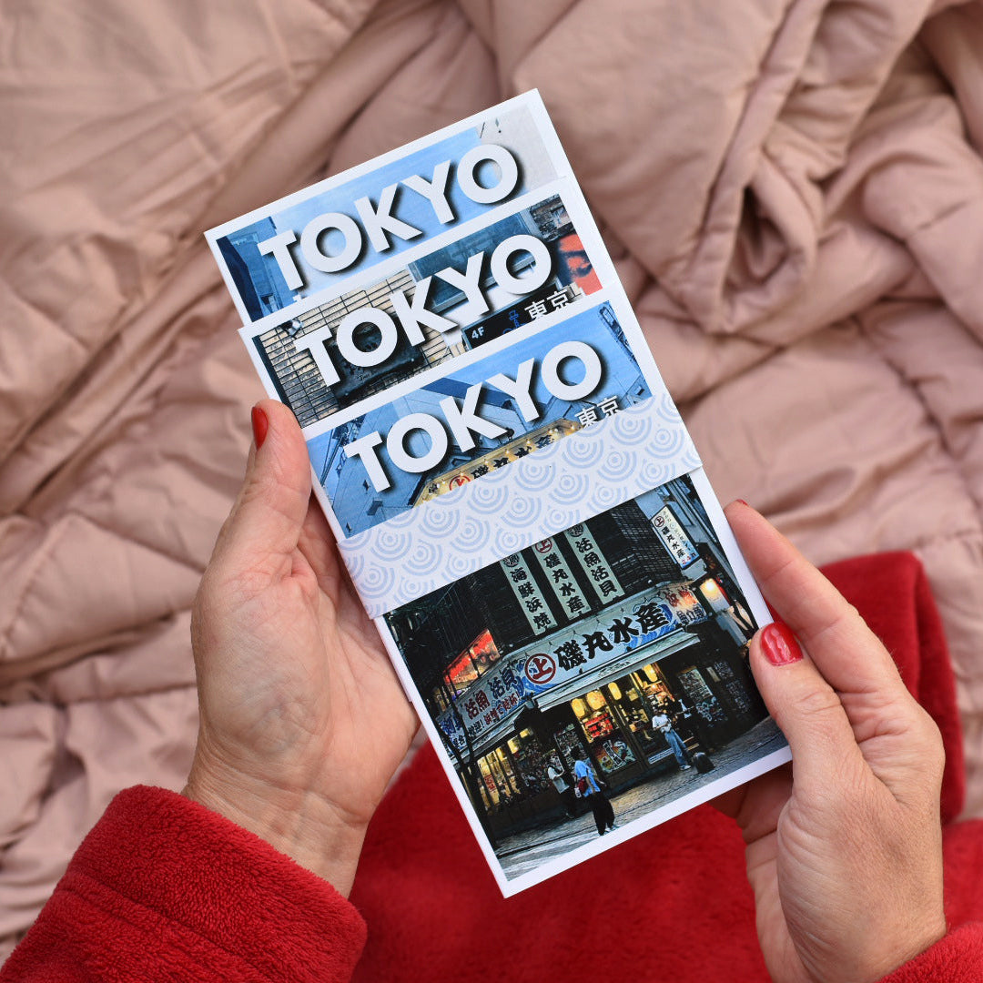 A person holds 3 Tokyo pocket travel guides featuring the word 'TOKYO' white, with a street scene photo at the bottom, against a pink quilted blanket background.