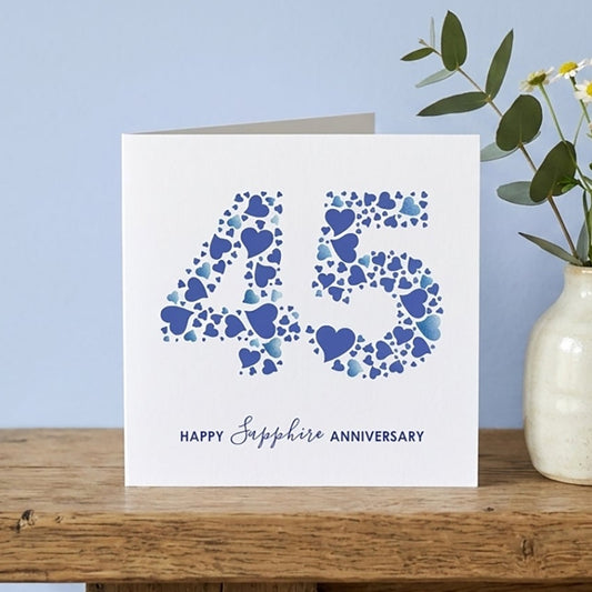 A square white greeting card with a blue '45' made of small hearts and the words 'HAPPY Sapphire Anniversary' sits on a rustic wooden table next to a small cream vase with flowers, against a light blue wall.