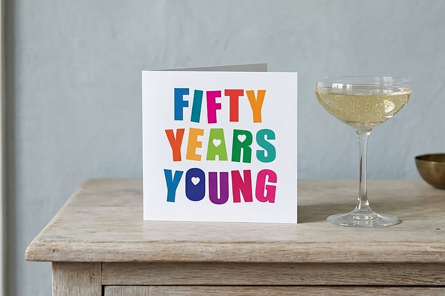 Card with 'Fifty Years Young' text on a wooden surface with a glass of champagne to celebrate.