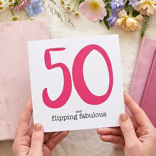 Person holding a card with '50 flipping fabulous' surrounded by notebooks and flowers on a light surface.