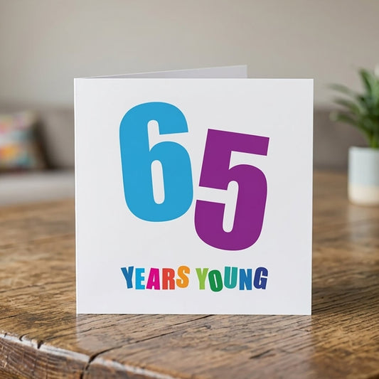 A white greeting card standing on a rustic wooden table, featuring the text '65 YEARS YOUNG' in colourful, playful lettering. The background shows a softly blurred living room setting with a sofa, a small potted plant, and a bookshelf.