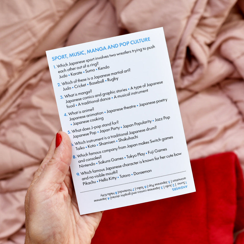 A hand holding a printed white card with blue text titled 'SPORT, MUSIC, MANGA AND POP CULTURE' against a soft pink blanket background.