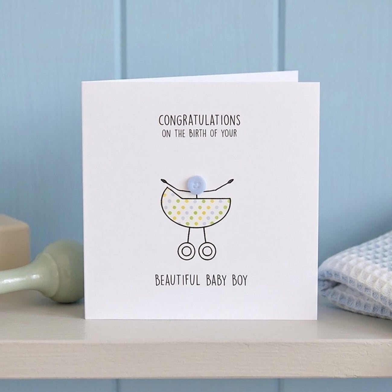 Baby shower card with baby pram illustration with a button baby on a shelf with toys against a blue wall.