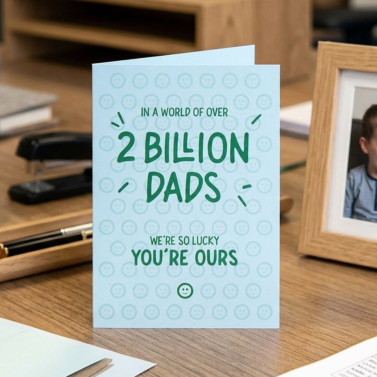 Card with text 'In a world of over 2 billion dads, we're so lucky you're ours' on a desk with a photo frame.