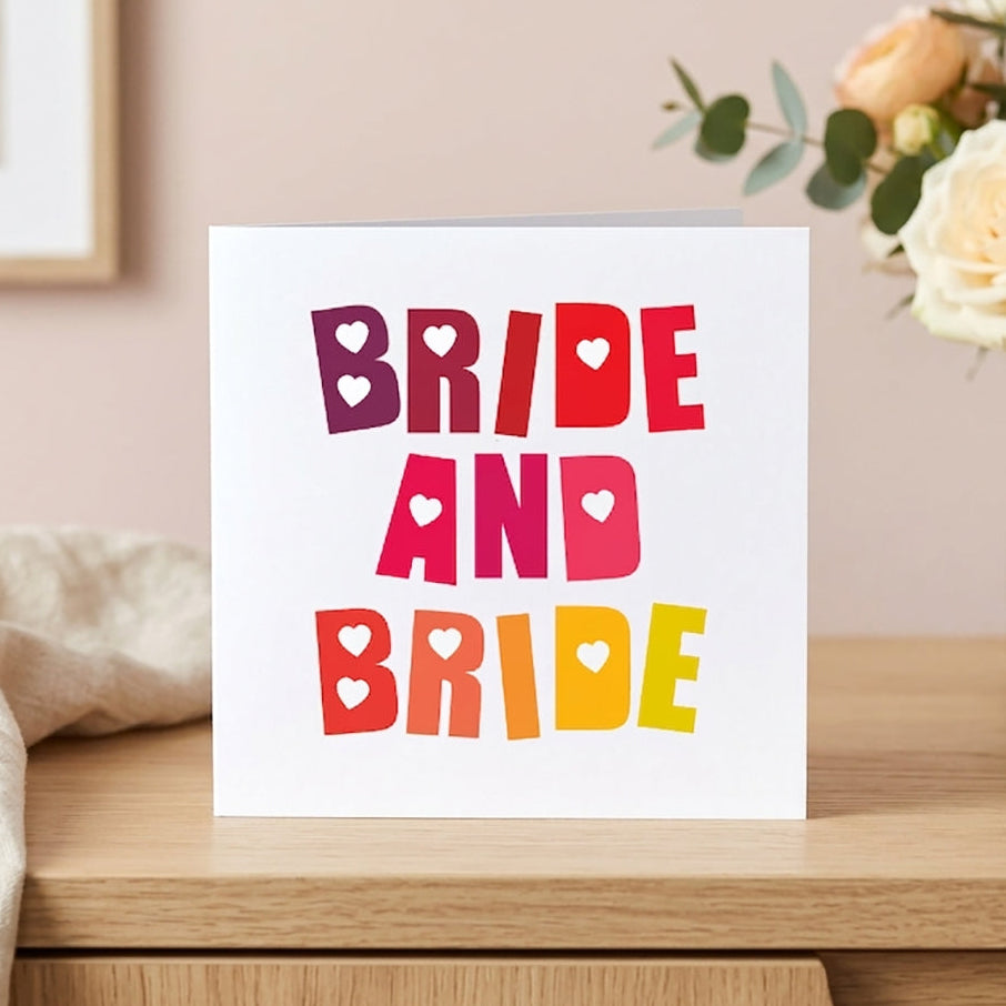 A vibrant bride and bride wedding card featuring a bold rainbow colour gradient typography layout with love hearts, styled beautifully in a bright home setting.