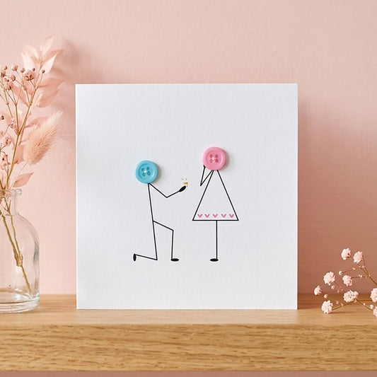 Handcrafted 3D button engagement card displayed on an oak shelf against a pale pink wall next to a small glass vase of pink flowers.