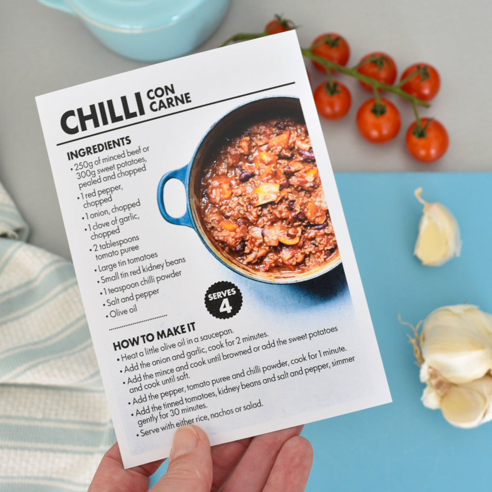 A hand holds a printed recipe card for Chilli Con Carne, styled on a light gray surface with a blue cutting board, garlic, tomatoes, and kitchen textiles around it.