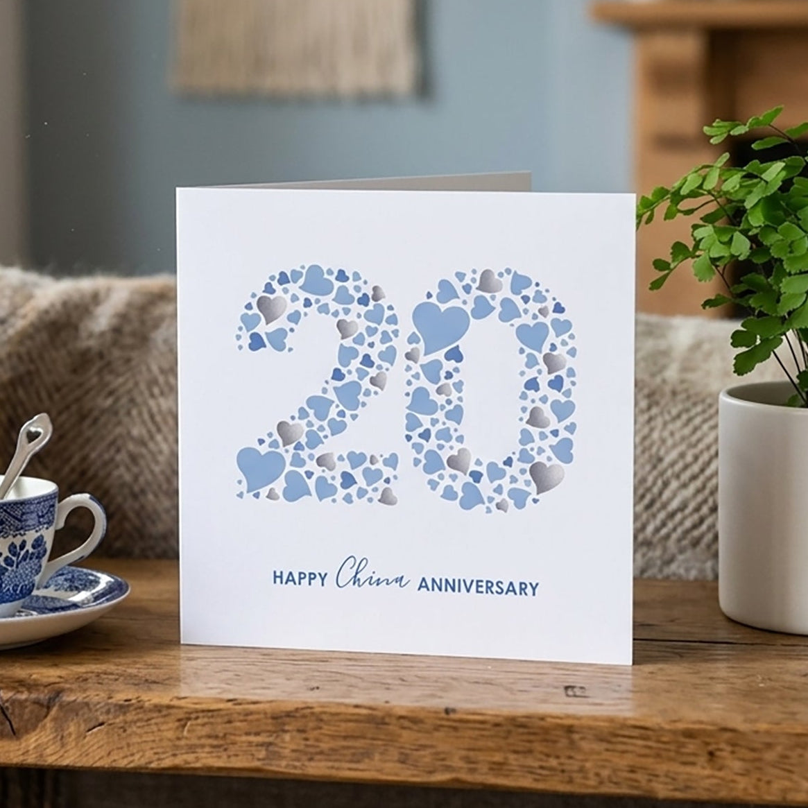 A gender-neutral 20th anniversary card for a husband or wife featuring the blue heart typography layout, displayed flat on a wooden table alongside books and a teacup.