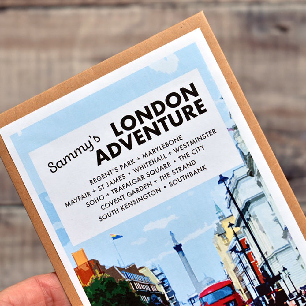 A hand holding a printed travel-style card or poster titled "Sammy's London Adventure" against a blurred wooden background. The card has a kraft-paper border, a light blue sky graphic, black text listing London landmarks, and a colourful illustration of red double-decker buses and city buildings along the bottom.