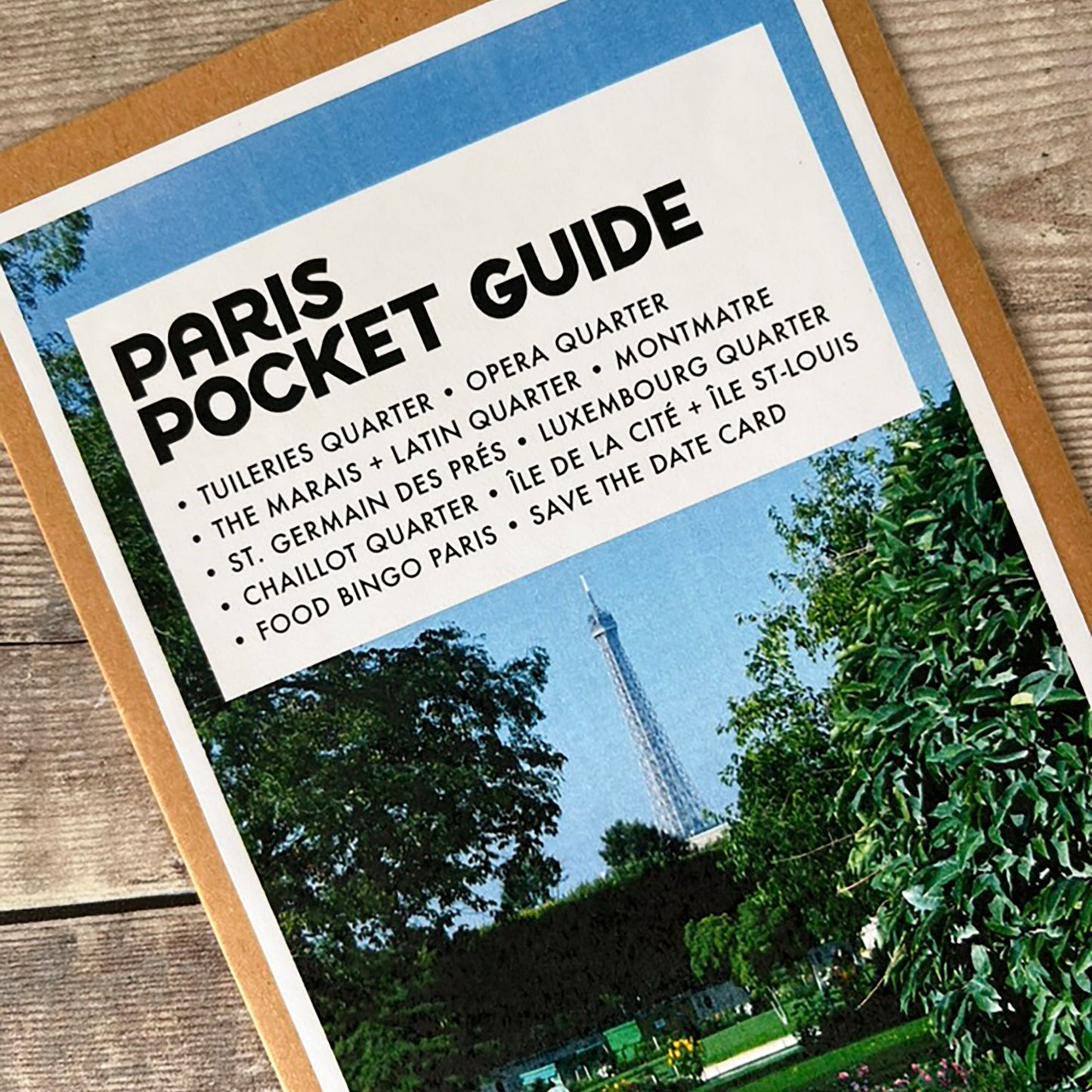 A Paris Pocket Guide pocket guide card with a kraft paper border, displayed diagonally on a rustic wooden surface. The card features a white header with black text listing various Parisian neighbourhoods and a scenic photograph of the Eiffel Tower in the background.