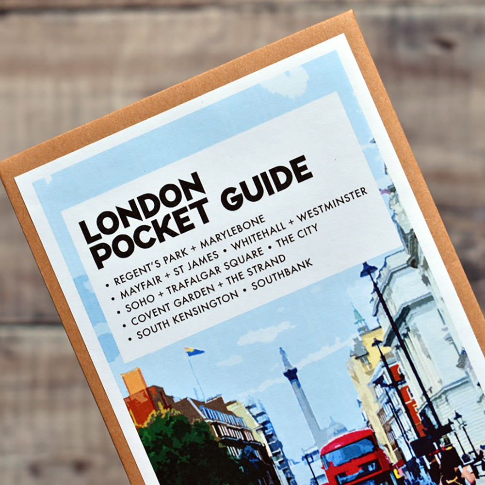 A physical copy of the 'London Pocket Guide' book shown at an angle against a blurred wooden background. The cover features a white top section with black text listing various London areas, and a bottom section with a colourful street scene illustration.