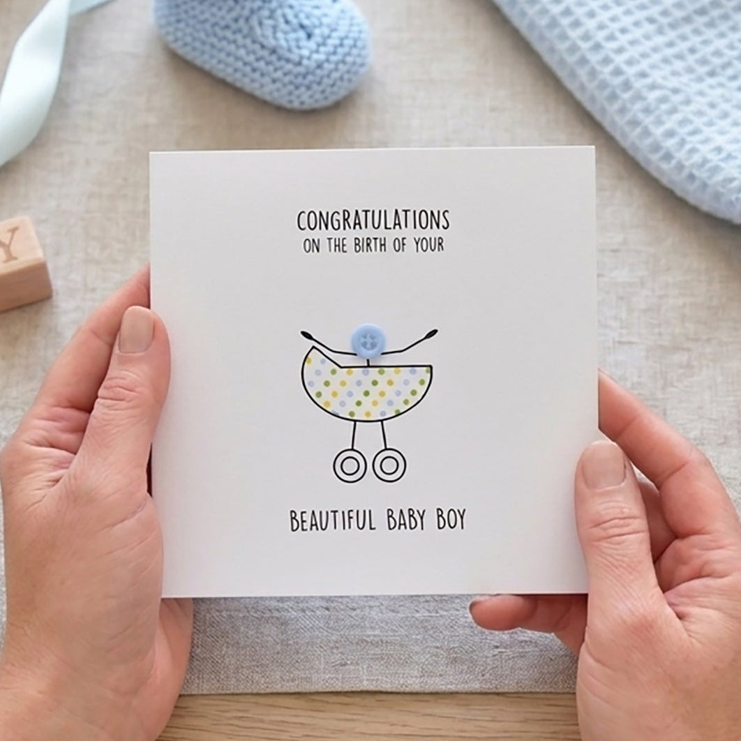 Person holding a new baby  card which reads "Congratulations on the birth of your beautiful baby boy" with baby booties, blanket and blocks on a table.
