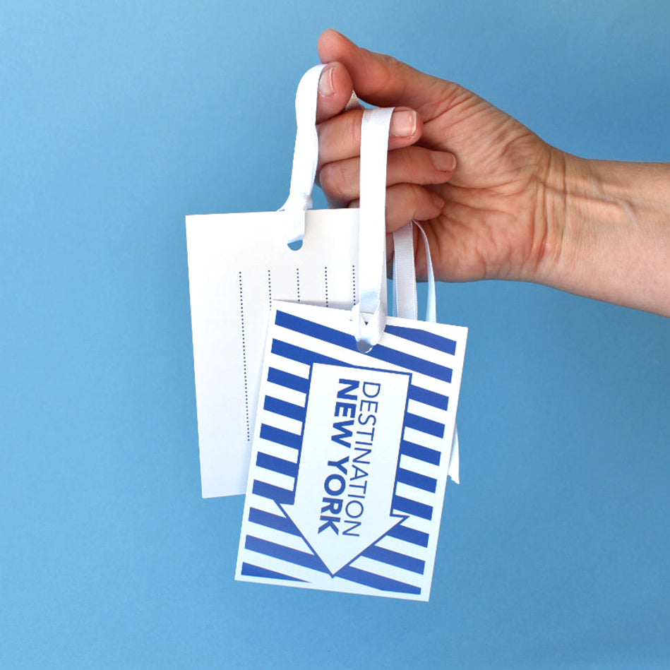 A hand holding a set of white luggage tags with blue stripes and the text 'DESTINATION NEW YORK' against a solid blue background.