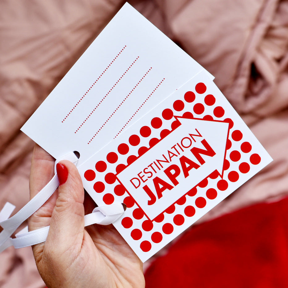 A hand holding a white luggage tags printed with red polka dots and the words 'DESTINATION JAPAN' inside an arrow, set against a soft pink blanket background.