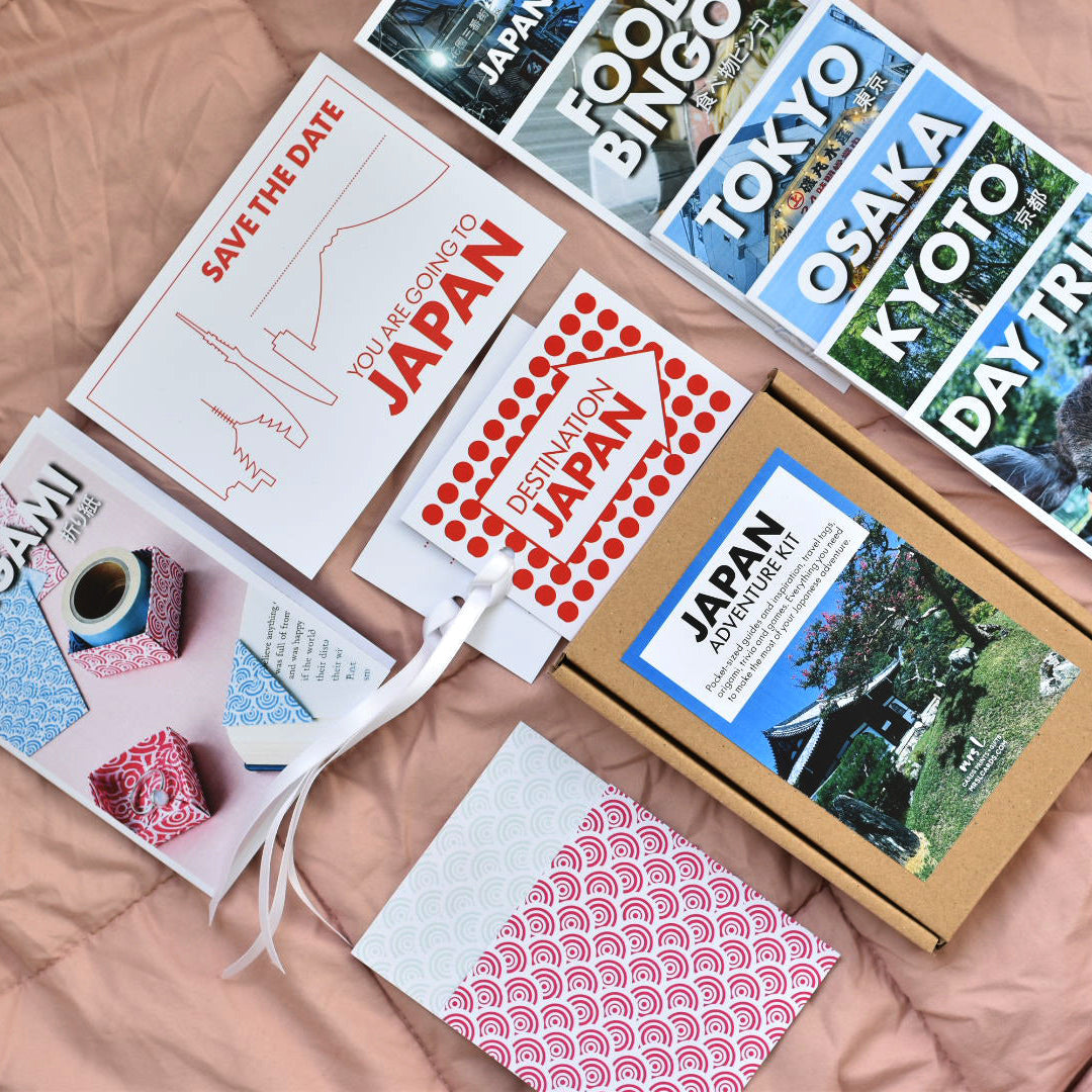 A curated flat-lay of the Japan Adventure Kit travel guide and gift items arranged on a pink quilted blanket, including printed cards, a patterned luggage ttag, a kraft box with a travel label and a small wrapped gift.