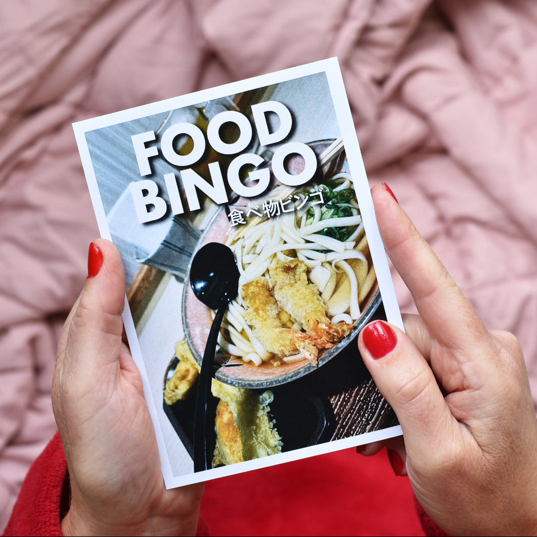A person holds a 'Food Bingo' card featuring a bowl of ramen, with their hands showing red nail polish against a pink quilted blanket background.