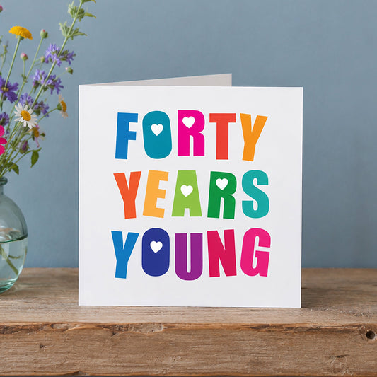 A white greeting card with the phrase 'FORTY YEARS YOUNG' in colorful retro letters sits on a rustic wooden shelf next to a glass vase of wildflowers against a muted blue-gray wall.