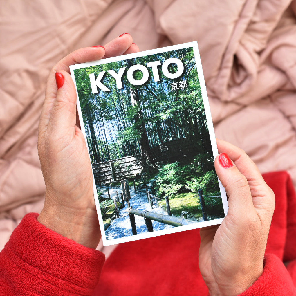 A person holding a pocket sized travel guide booklet titled "KYOTO" in front of their chest. The cover shows a calm garden scene with a bridge over water.