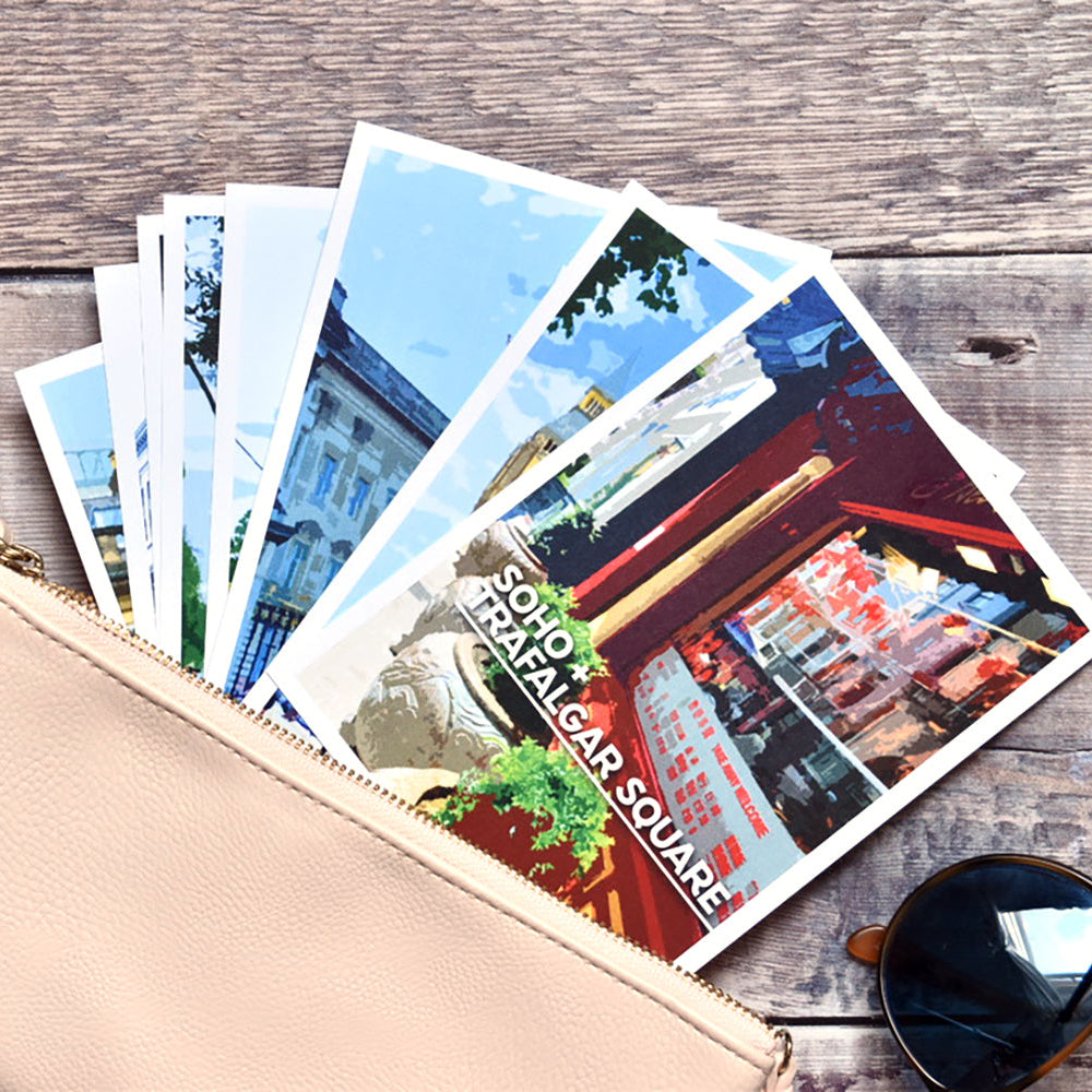 A beige zip pouch with a wrist strap lies open on a rustic wooden surface, fanning out several postcards. One postcard shows a street scene with the text 'SOHO + TRAFALGAR SQUARE".