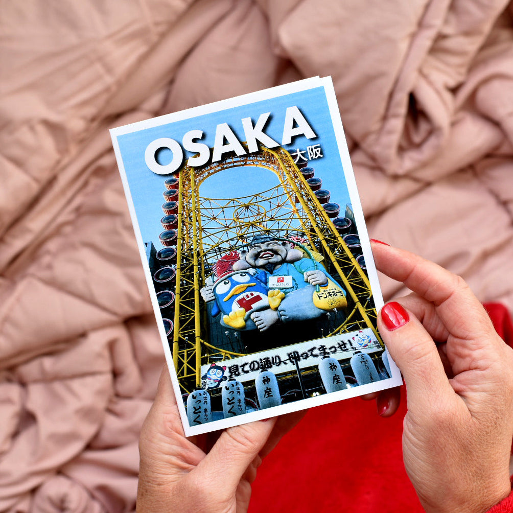 A person holds a rectangular Osaka travel postcard featuring a yellow roller coaster and the text 'OSAKA' against a soft pink quilted blanket background.