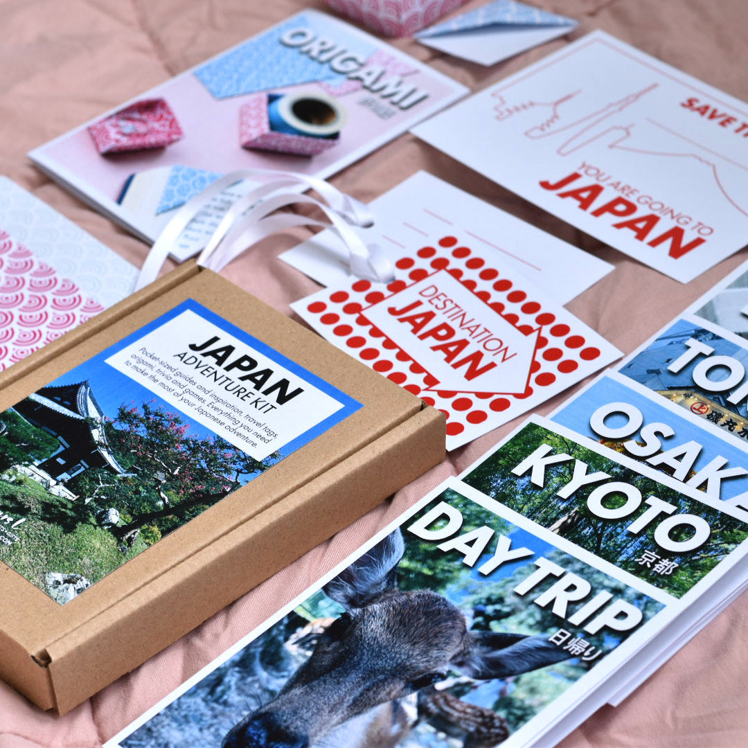 A curated collection of Japan-themed guides and gift items arranged on a pink fabric background, including a brown cardboard box with a Japan picture cover a white origami booklet, a red 'Save the Date' card, pink origami paper and a blue-and-white 'Day Trip' itinerary card.