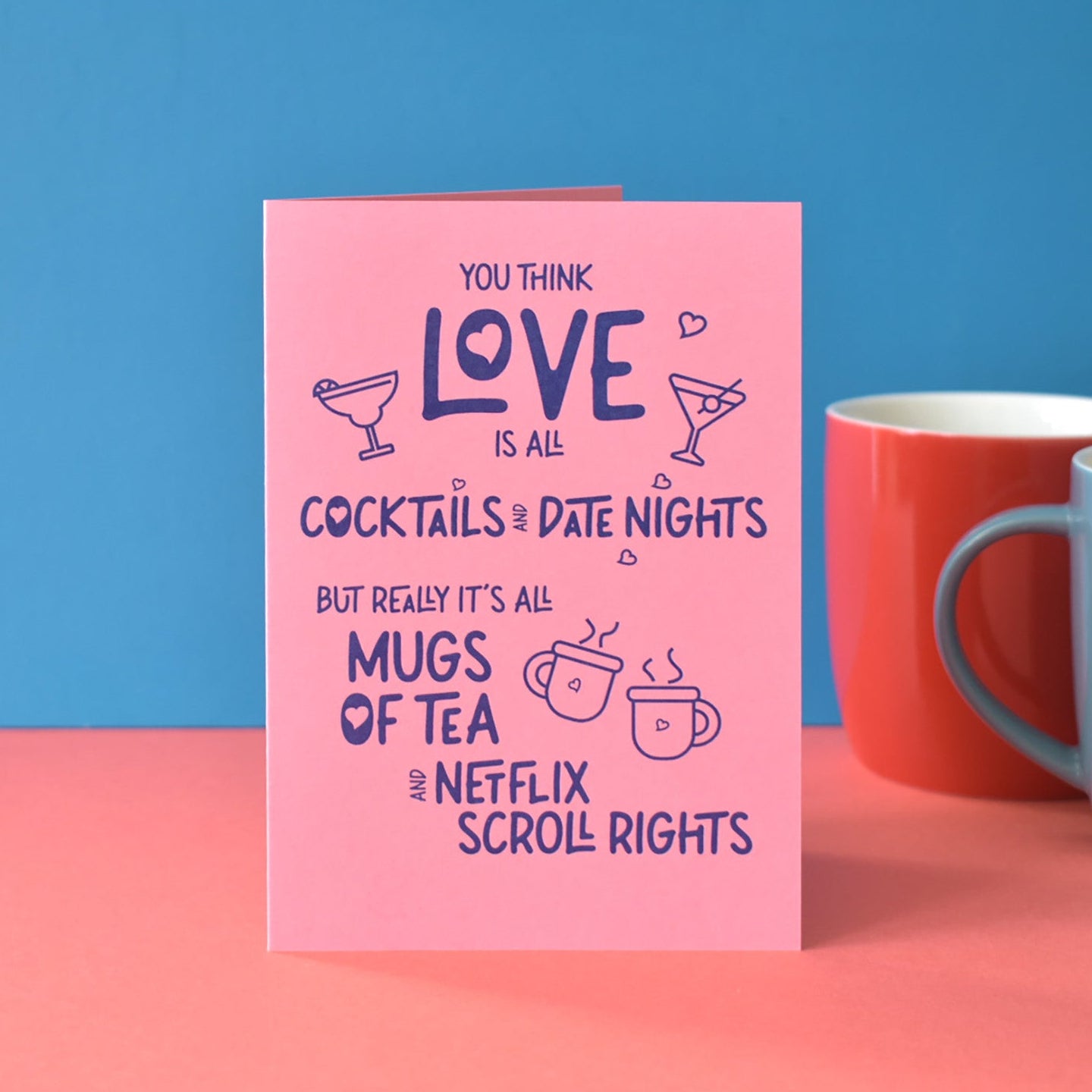 Pink card with humorous text about love and mugs of tea, against a blue and pink background with two mugs. Text reads ""You think love is all cocktails and fancy date nights, but really it's all mugs of tea and Netflix scroll rights."