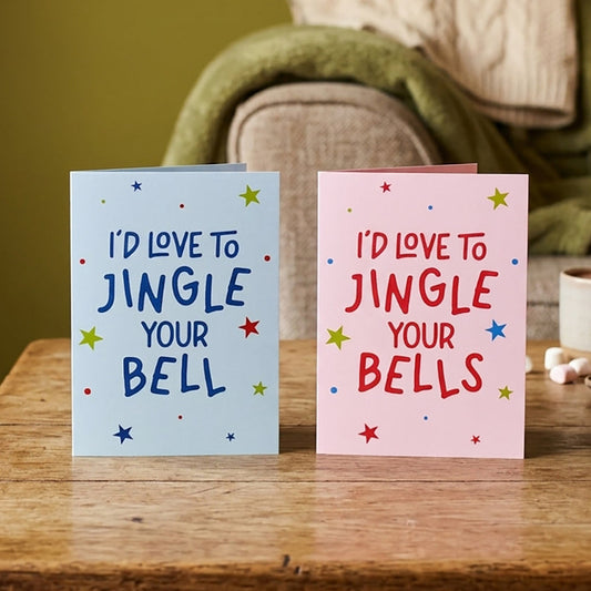 Two Christmas greeting cards standing on a wooden table in a cozy living room setting. One card is light blue with dark blue text reading 'I'D LOVE TO JINGLE YOUR BELL' and small multicolored stars and confetti.