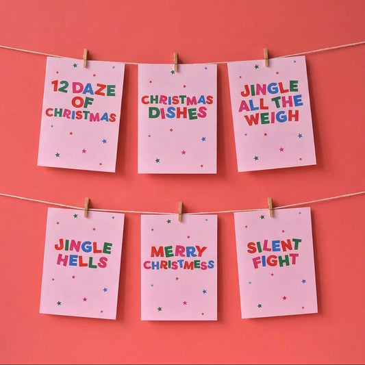 Six pink Christmas greeting cards with colorful text hang from two strings on a coral pink background. The cards read: '12 DAZE OF CHRISTMAS', 'CHRISTMAS DISHES', 'JINGLE ALL THE WEIGH', 'JINGLE HELLs', 'MERRY CHRISTSS', and 'SILENT FIGHT'.