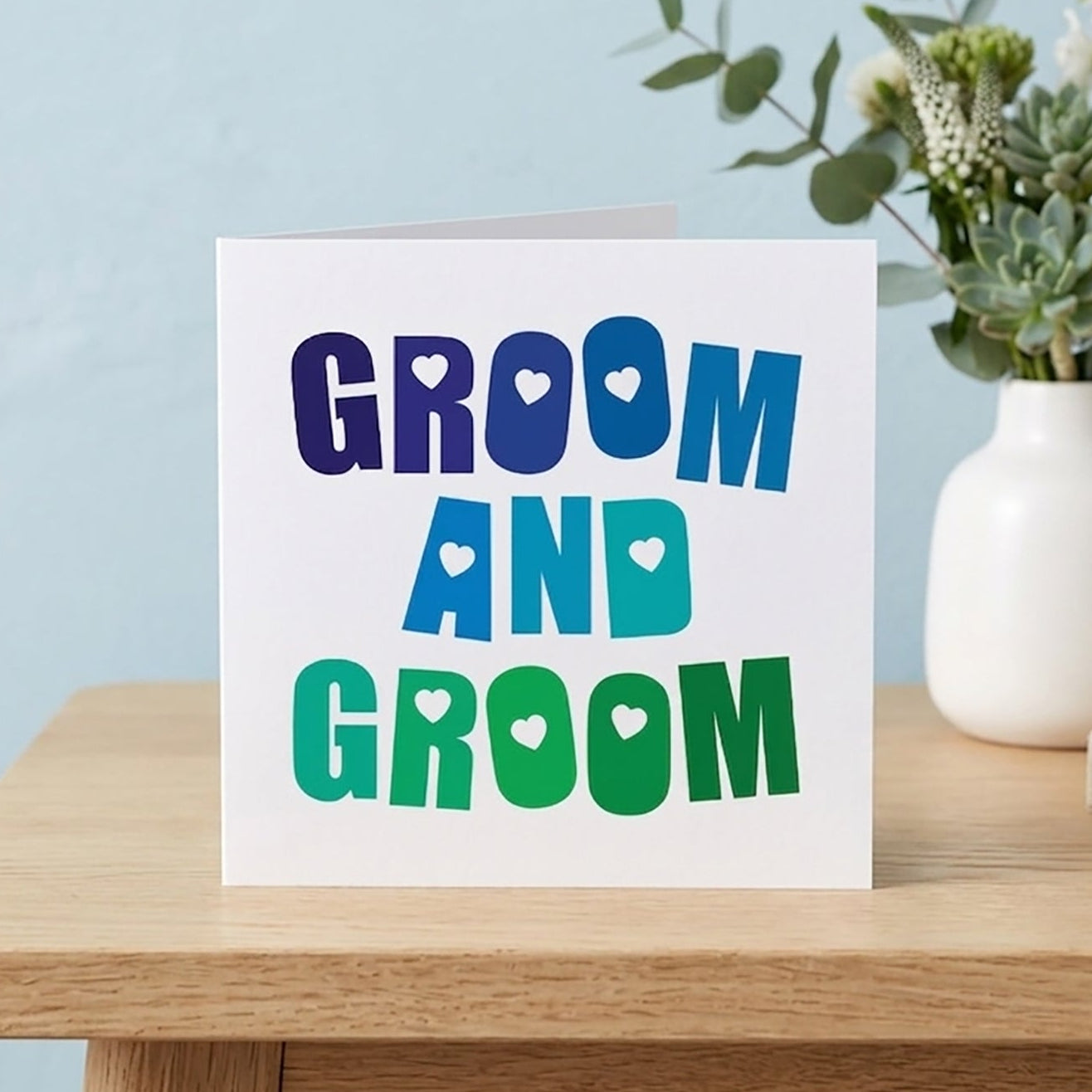 A vibrant groom and groom wedding card featuring a bold blue and green rainbow typography layout with love hearts, perfect for a gay male wedding.