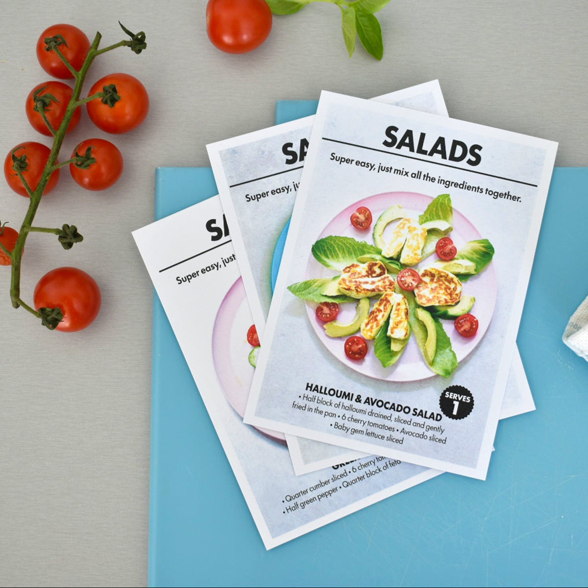 Three printed recipe cards for salads are stacked on a blue folder, styled with fresh tomatoes, basil, and a striped cloth on a light gray surface.