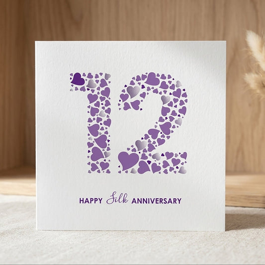 A square white 12th Silk Anniversary card by mrs L cards, featuring a large number 12 filled with a pattern of purple and silver hearts. Below the number, clean typography reads 'Happy Silk Anniversary' in a deep purple font.
