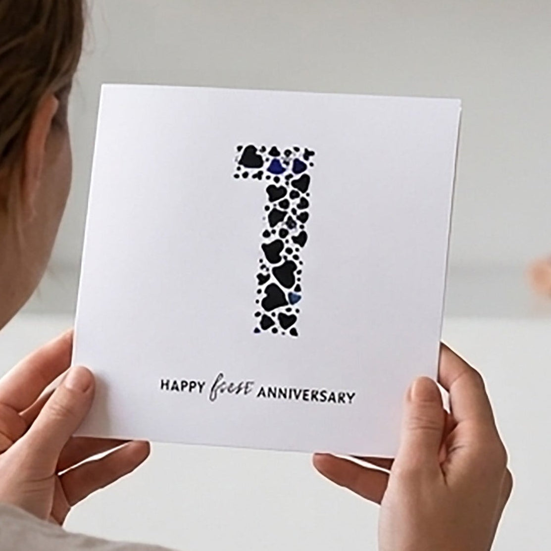 Couple sitting on a couch holding a first wedding anniversary card with a large number 1 made from a collection of love hearts and the words Happy First Anniversary in text underneath the number 1.  The couple are sat in a cozy living room setting.