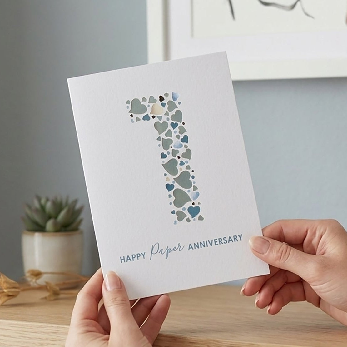 Person holding a card with a large number '1' and 'Happy Paper Anniversary' text, sitting on a wooden shelf.
