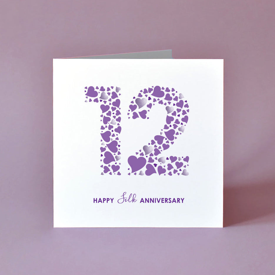 A stylishly simple 145mm square Silk Anniversary card standing upright. The design shows a colourful mosaic of purple and grey hearts forming the number 12 on a crisp white background, styled next to its matching white envelope.