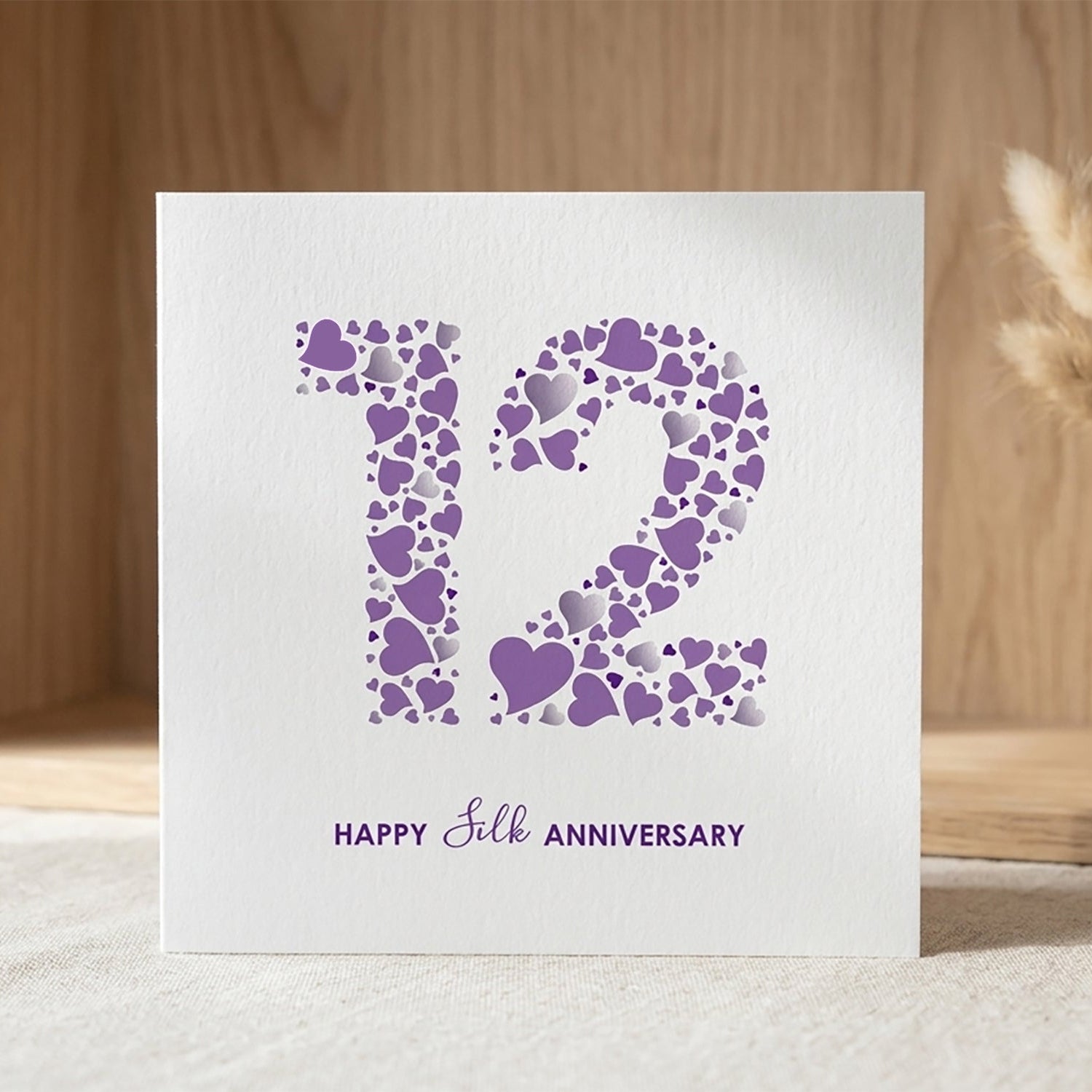 A square white 12th Silk Anniversary card by mrs L cards, featuring a large number 12 filled with a pattern of purple and silver hearts. Below the number, clean typography reads 'Happy Silk Anniversary' in a deep purple font.