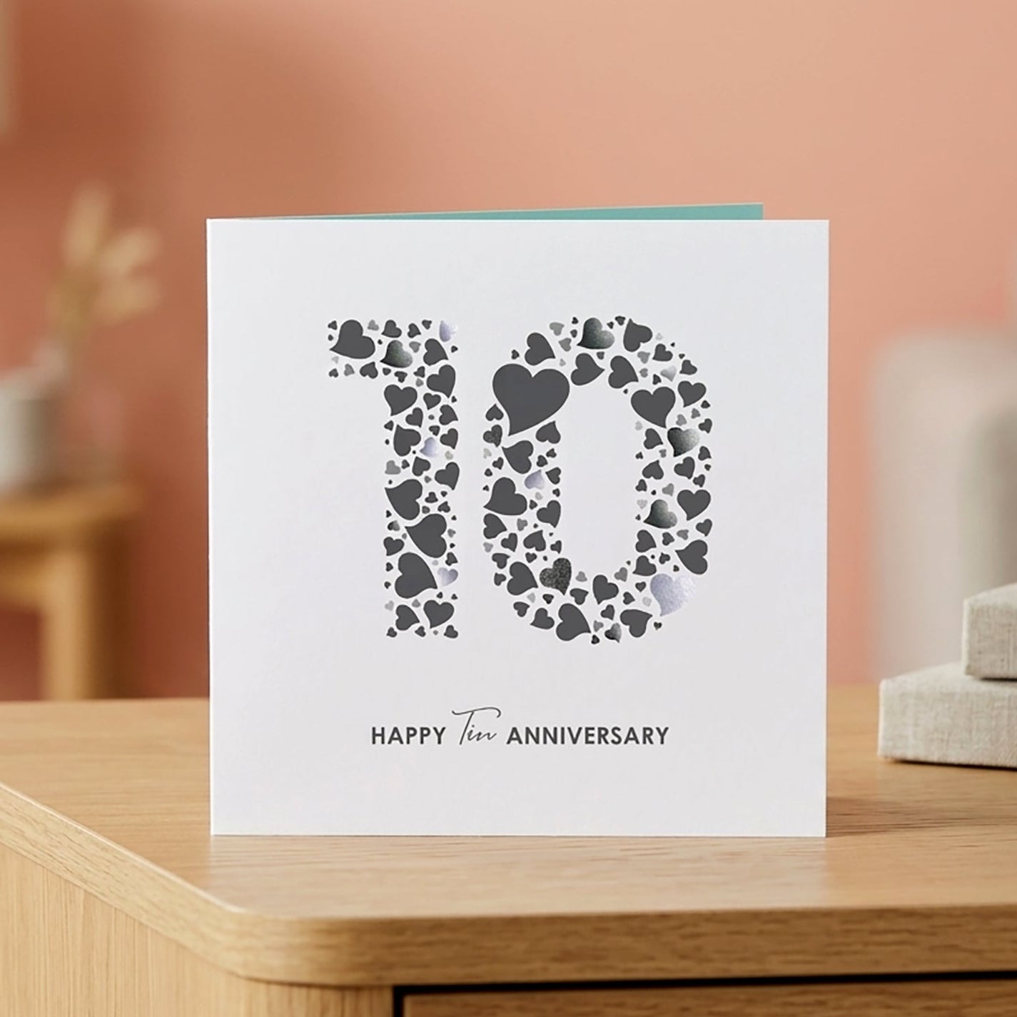 A square white 10th wedding anniversary card with a modern "10" design made of grey and light grey love hearts, displayed on a wooden table in a living room with light coral walls and blurred neutral decor.