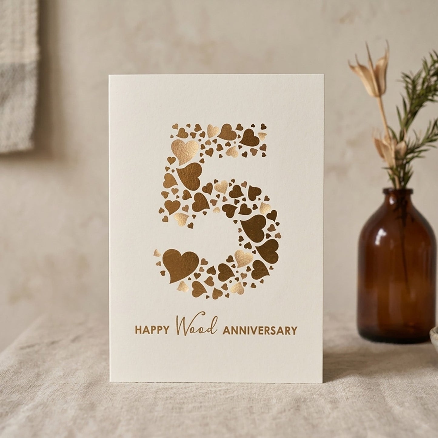 Front view of a 5th wedding anniversary card on smooth cream card stock with  brown hearts making up the number 5, displayed against a neutral plaster-coloured wall with soft natural fabrics.