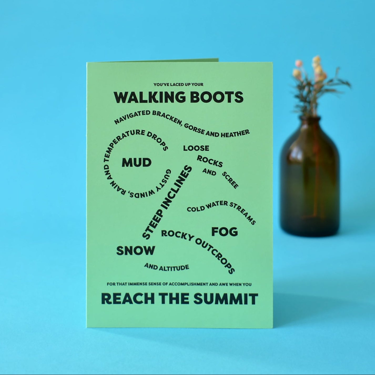 Green card with bold, black text that reads The wording on the card reads "You've laced up your walking boots, navigated bracken, gorse and heather, loose rocks and scree, gusty winds, rain and temperature drops, mud, steep inclines, cold water streams, fog, rocky outcrops, snow and altitude for that immense sense of accomplishment and awe when you reach the summit."