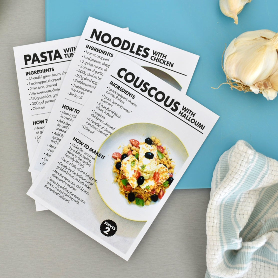Two printed recipe cards for pasta and couscous with chicken are laid on a gray surface, surrounded by a yellow spatula, garlic, a blue backdrop, and a striped cloth. The cards show ingredients, instructions, and a photo of the prepared dish.