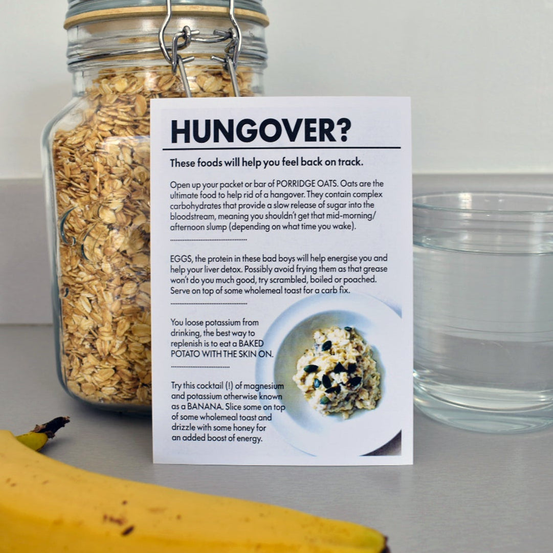A printed hangover recovery card titled 'HUNOVER?' placed on a countertop next to a glass of water, a jar of oats, and a banana. The card includes text and a small food photo.