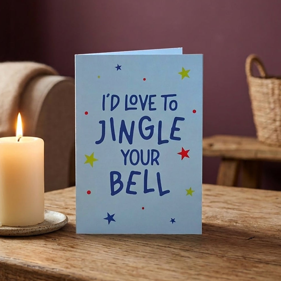 A light blue greeting card with the text 'I'D LOVE TO JINGLE YOUR BELL' in dark blue lettering, decorated with small colorful stars, sits on a rustic wooden table next to a lit cream-colored candle in a cozy living room setting.